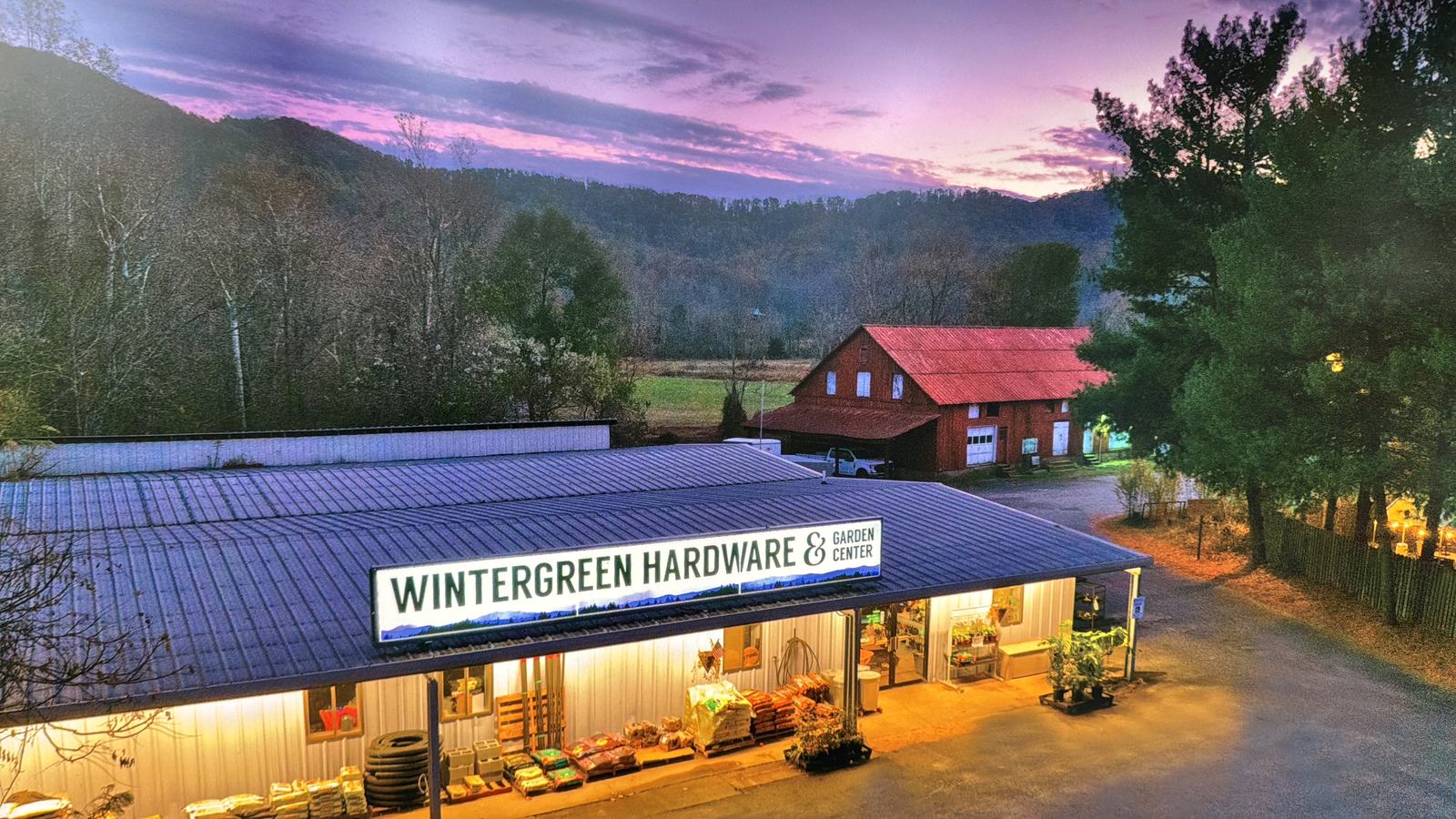 Wintergreen Hardware & Garden Center from the air at dusk: the long metal-roofed store with its lit sign, a red-roofed barn behind it, and wooded Blue Ridge hills under a pink and purple sky